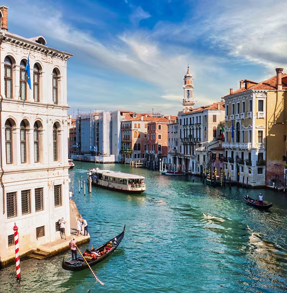Venetian canal with gondolas, a water bus, and historic buildings under a blue sky with scattered clouds.