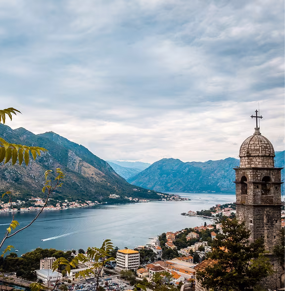 Scenic view of a coastal town with mountains, water bay, and a historic stone tower with a cross on top.