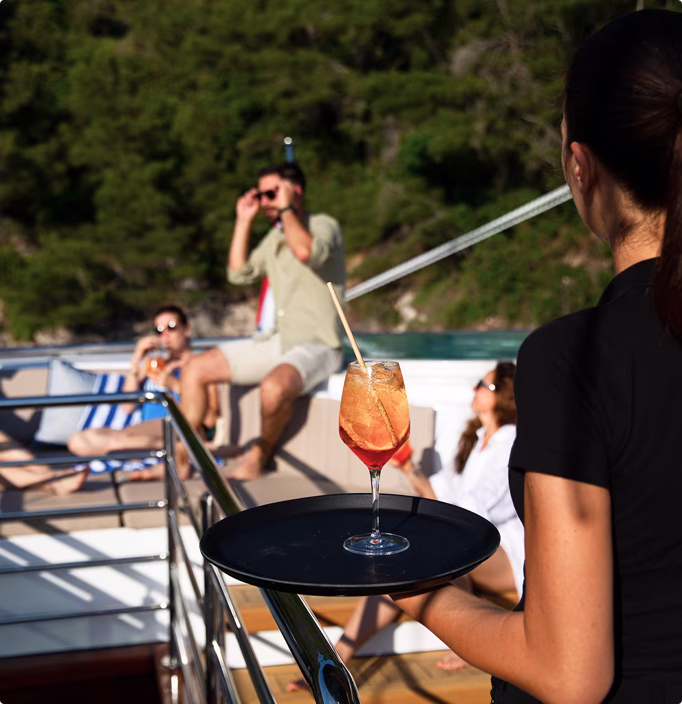Waitress holding a tray with a cocktail on a yacht, with people relaxing and wearing sunglasses in the background.