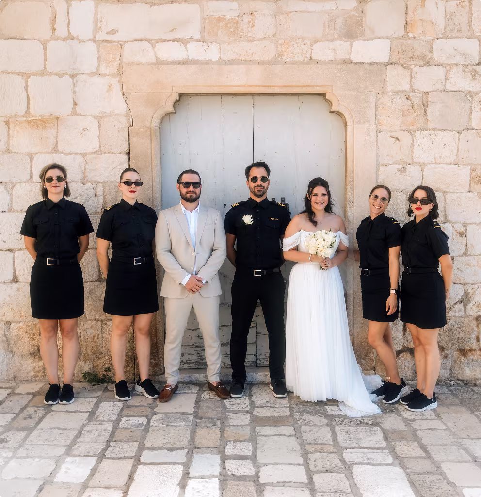 Bride and groom standing with four women and one man in black uniform dresses and black pants in front of a stone wall and wooden door.