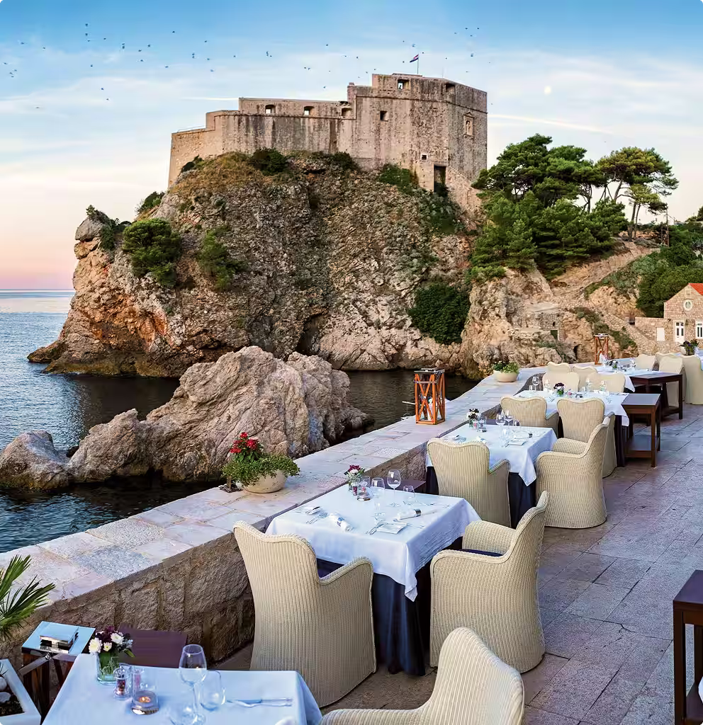 Outdoor restaurant terrace with wicker chairs and white tablecloths overlooking rocky cliffs and historic stone fortress by the sea.