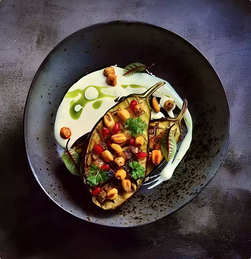 Grilled eggplant halves garnished with herbs, nuts, and pomegranate seeds on a black plate with a creamy white sauce and green oil drizzle.