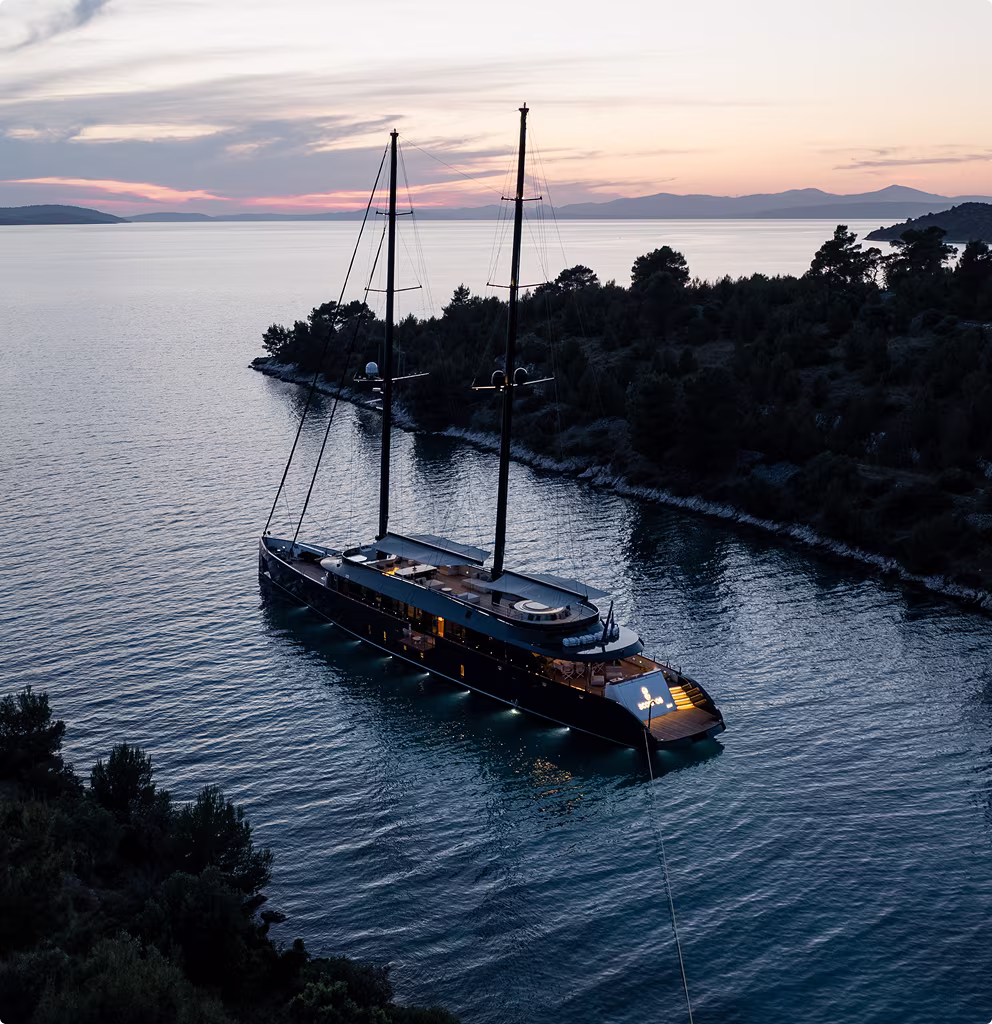 Large yacht anchored near a forested shoreline at dusk with calm waters and a colorful sky.