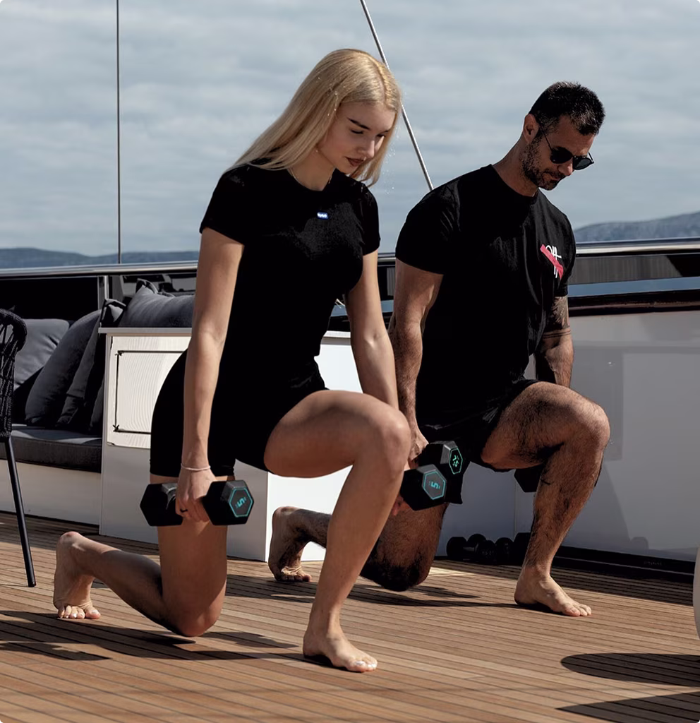 A man and a woman performing lunges while holding dumbbells on a wooden deck outdoors.