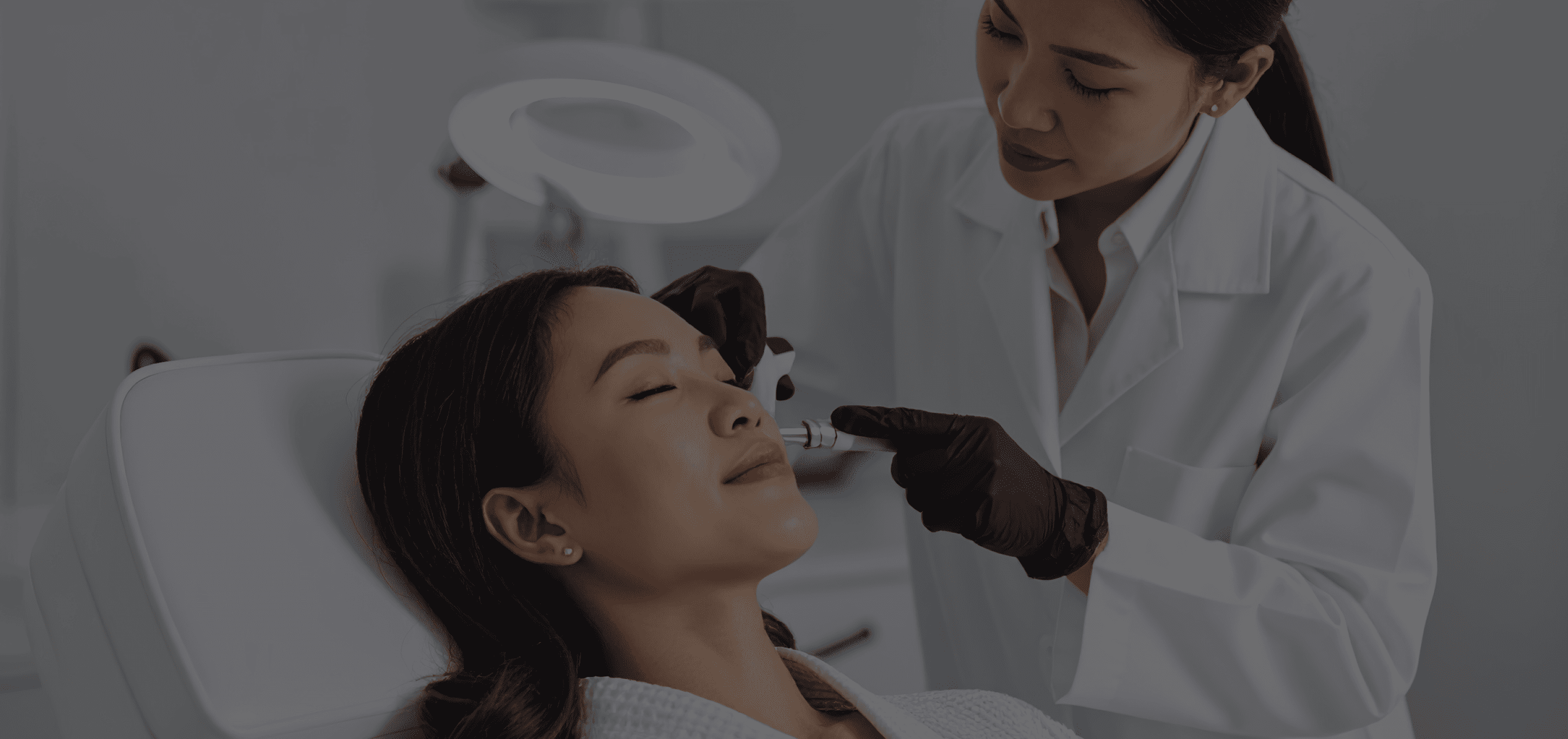 A cosmetologist wearing black gloves and white coat administers a facial treatment to a relaxed woman lying in a white chair.