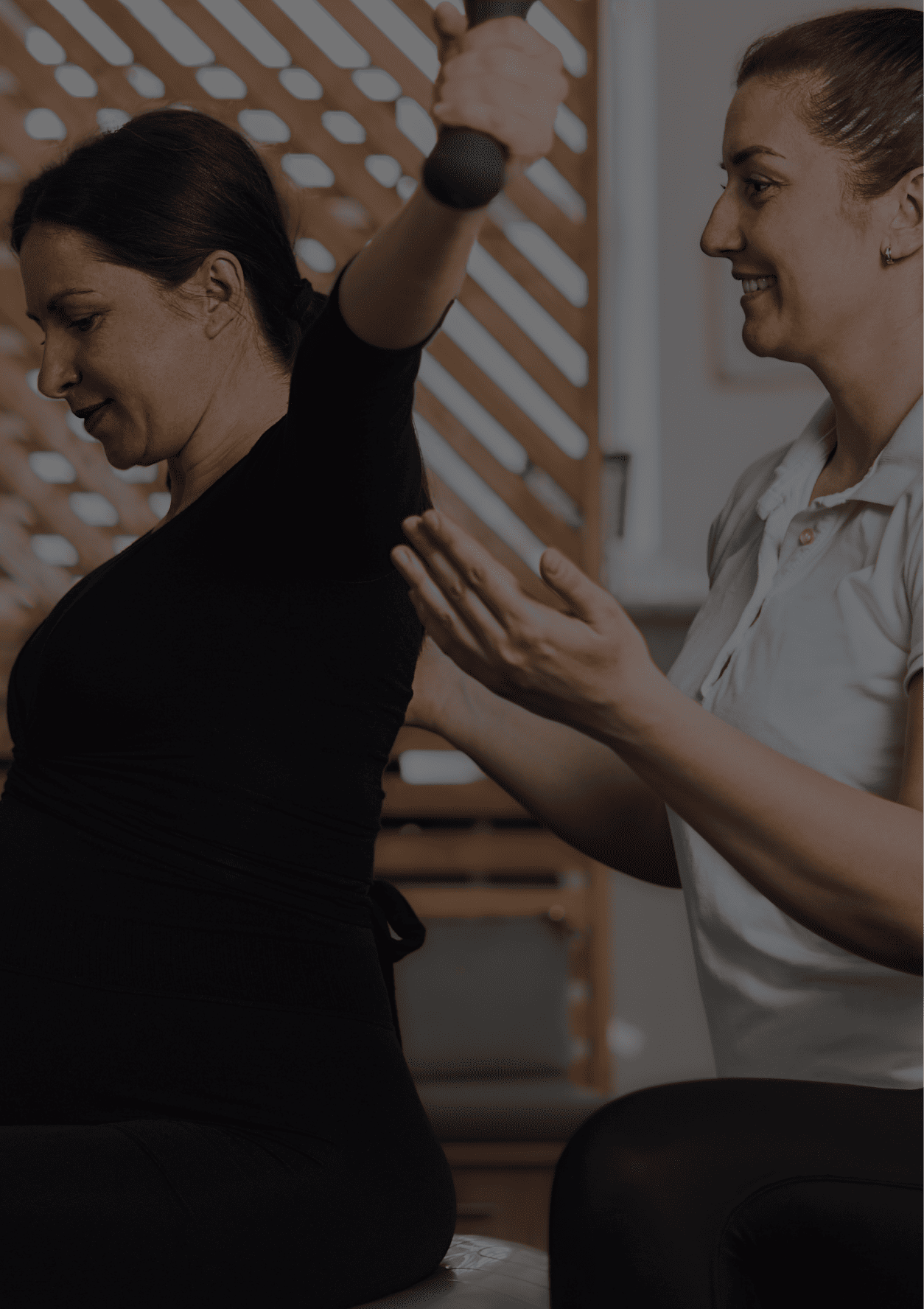 A woman in black lifts a bar while a trainer in white guides her posture with hands.