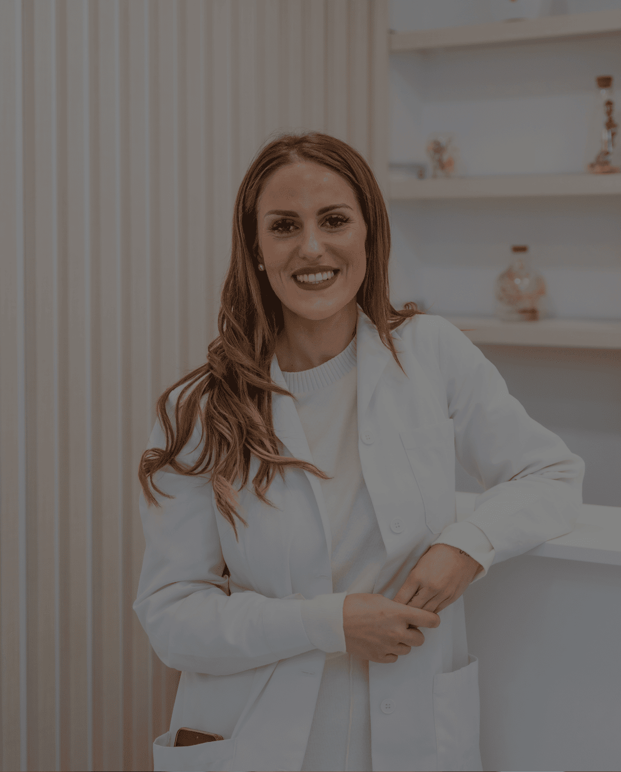 Smiling woman in a white medical coat standing with her hand in her coat pocket in a treatment or consultation room.