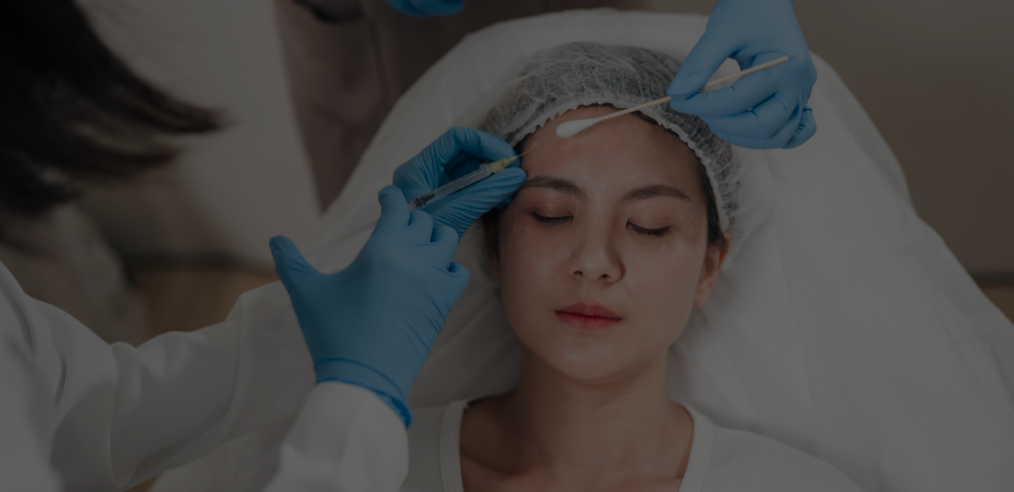 Woman with closed eyes receiving a cosmetic injection in the forehead by a person wearing blue gloves.