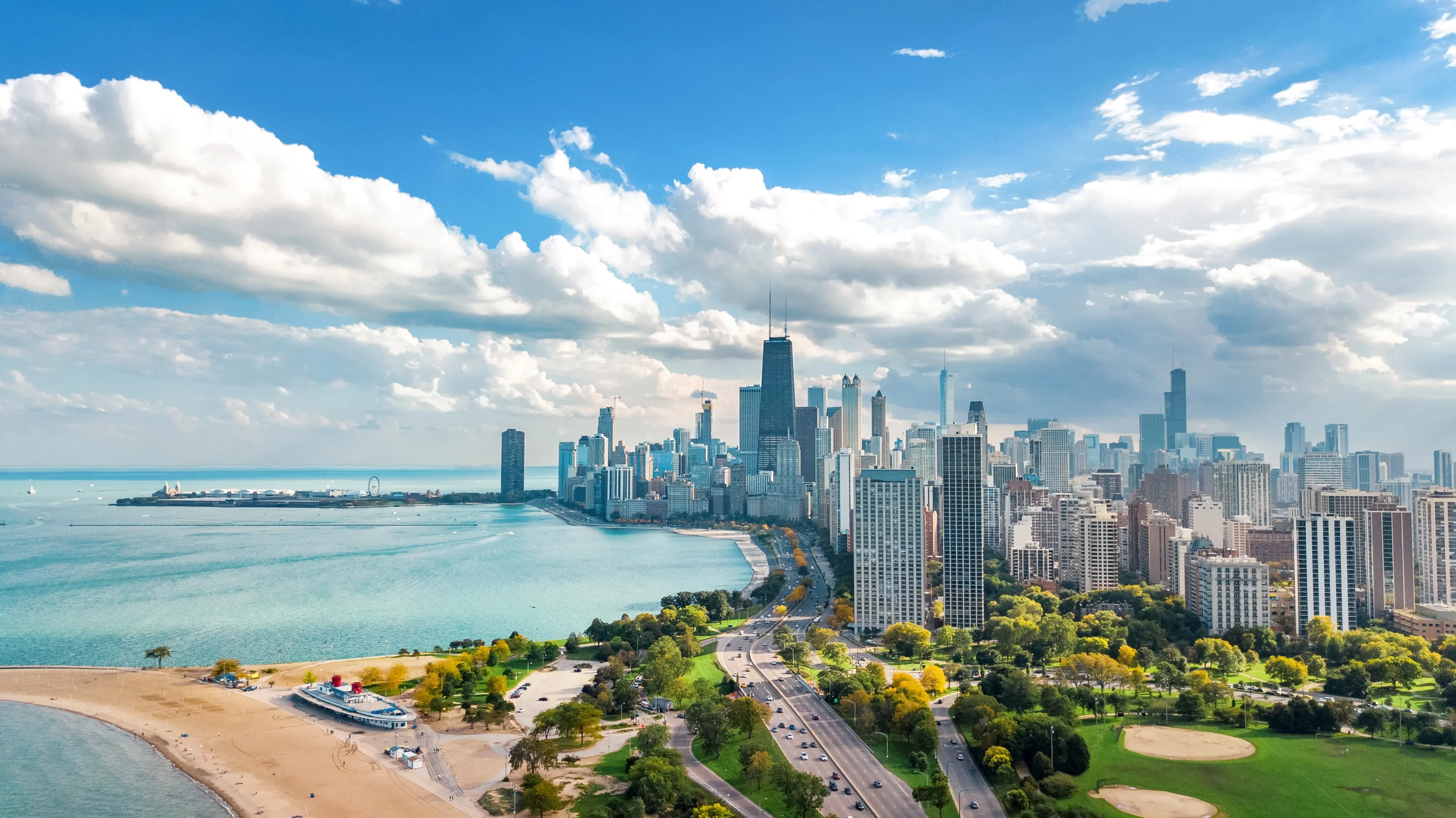 Aerial view of a city skyline with tall buildings along a lakefront, featuring a beach, green parks, and a partly cloudy blue sky.