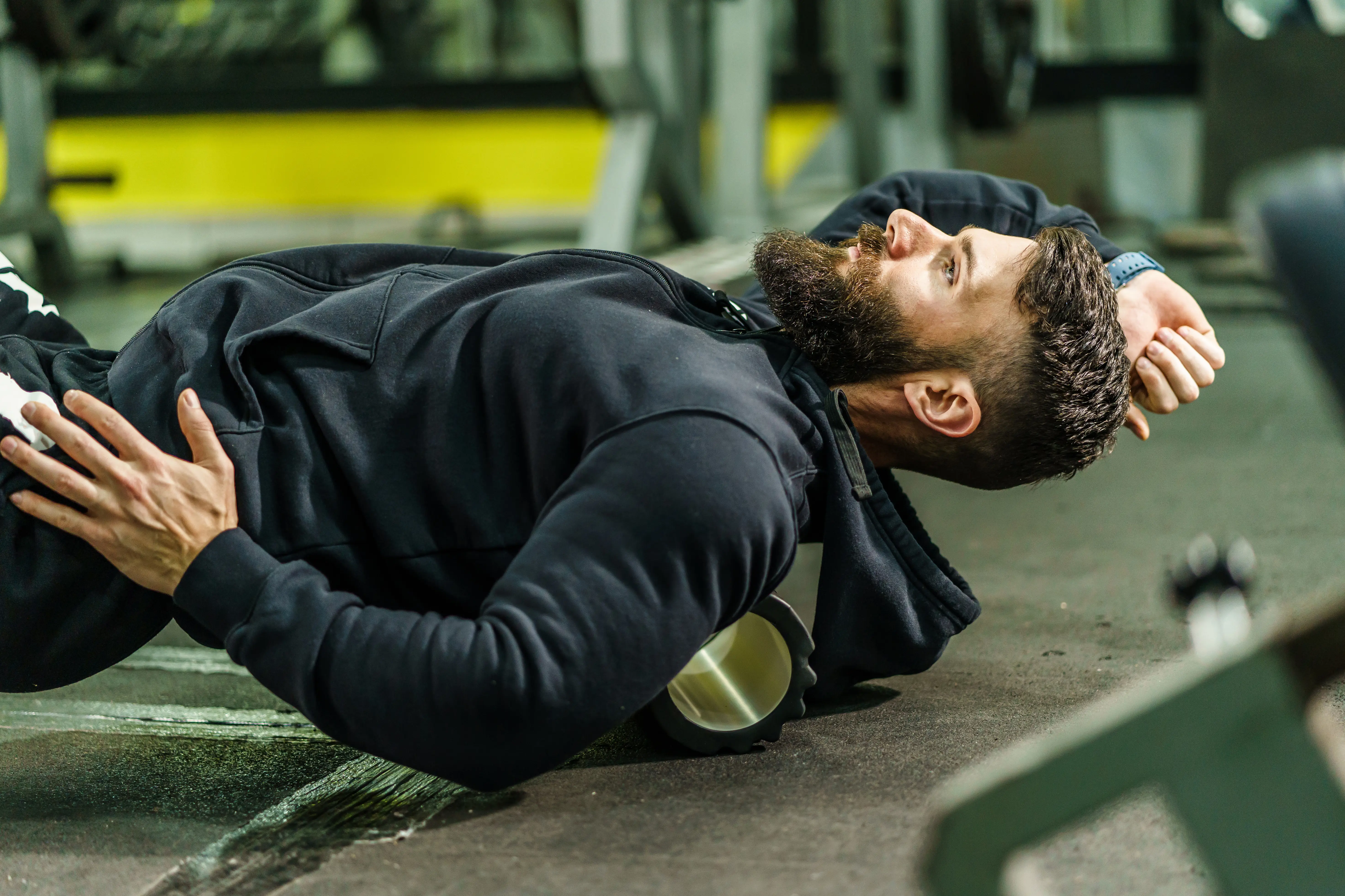 Bearded man in black hoodie using a foam roller on his upper back in a gym.
