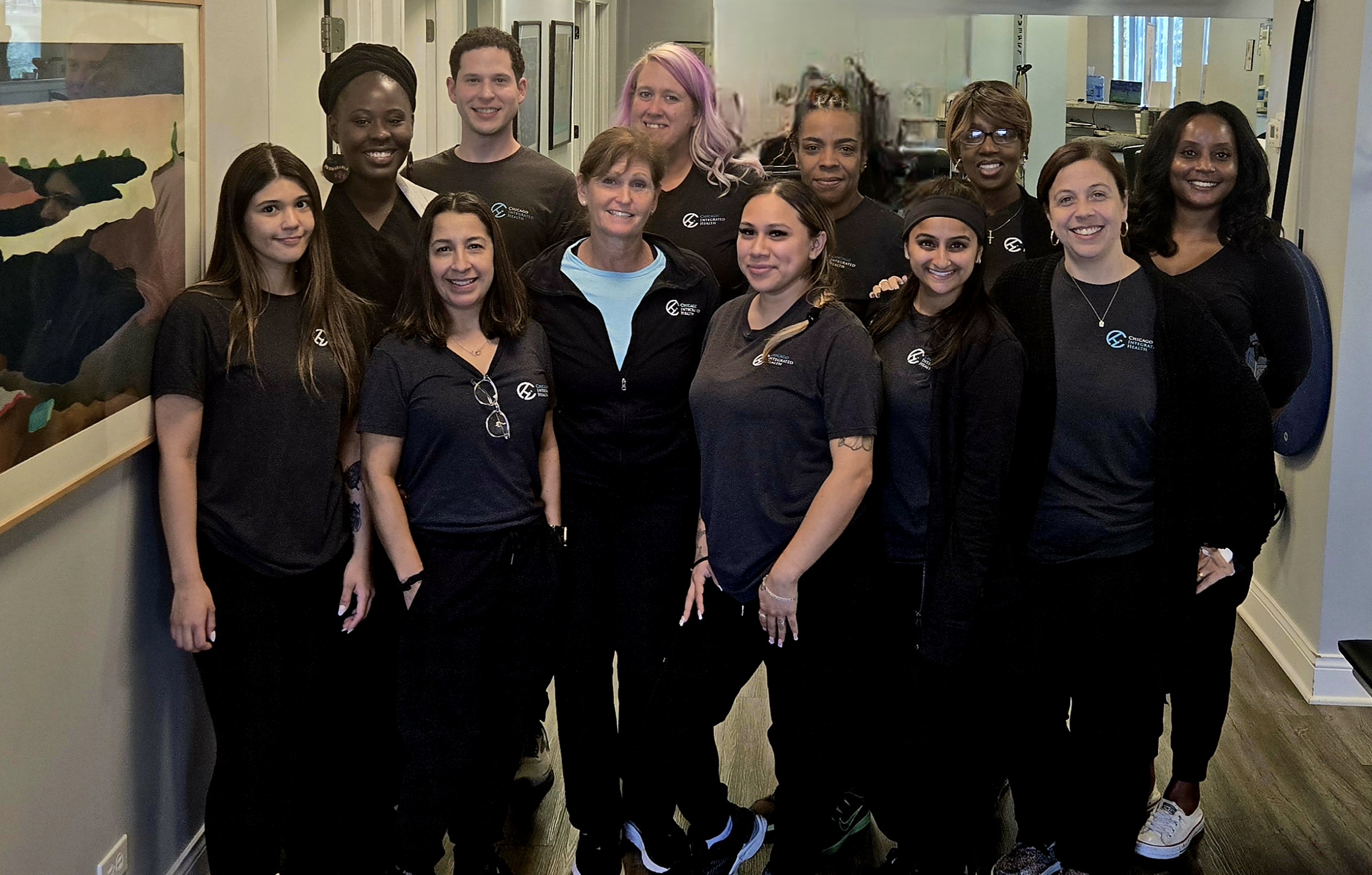 Group of nine smiling people standing together in a fitness or physical therapy center with exercise balls and equipment in the background.