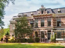 Groot statig gebouw van baksteen met grote ramen en een dak met houten details, omgeven door bomen en gras.