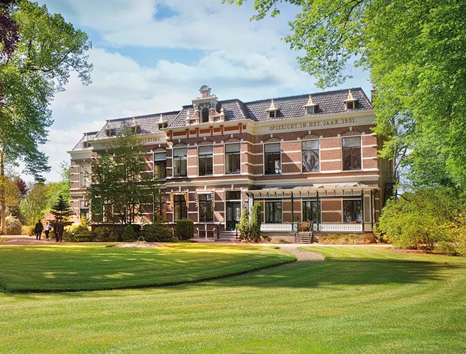 Groot historisch herenhuis uit 1901 met roodbruine bakstenen en wit sierwerk, omgeven door een groen gazon en bomen.