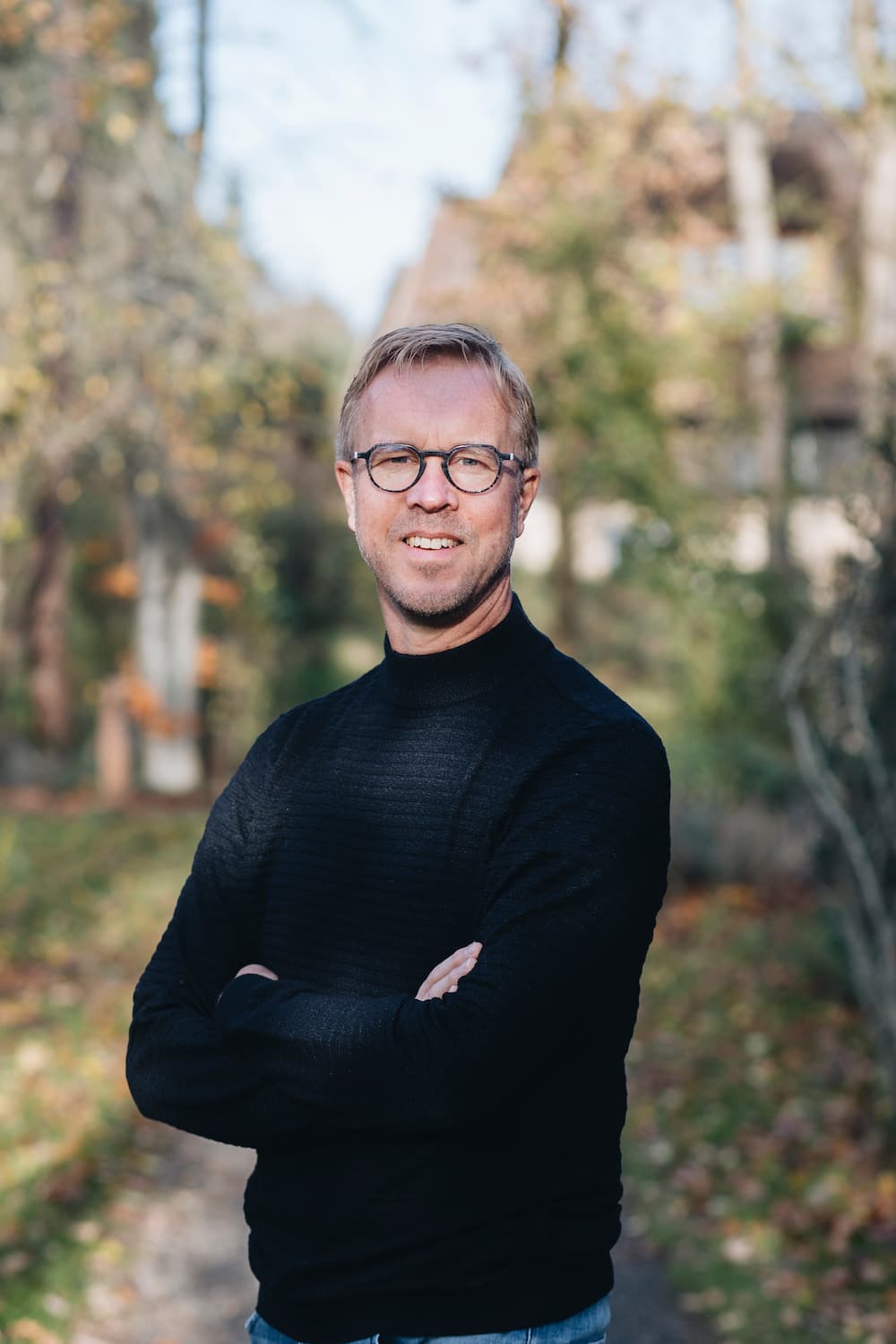 Man met bril en zwarte trui staat met gekruiste armen buiten op een pad omgeven door herfstachtig groen.