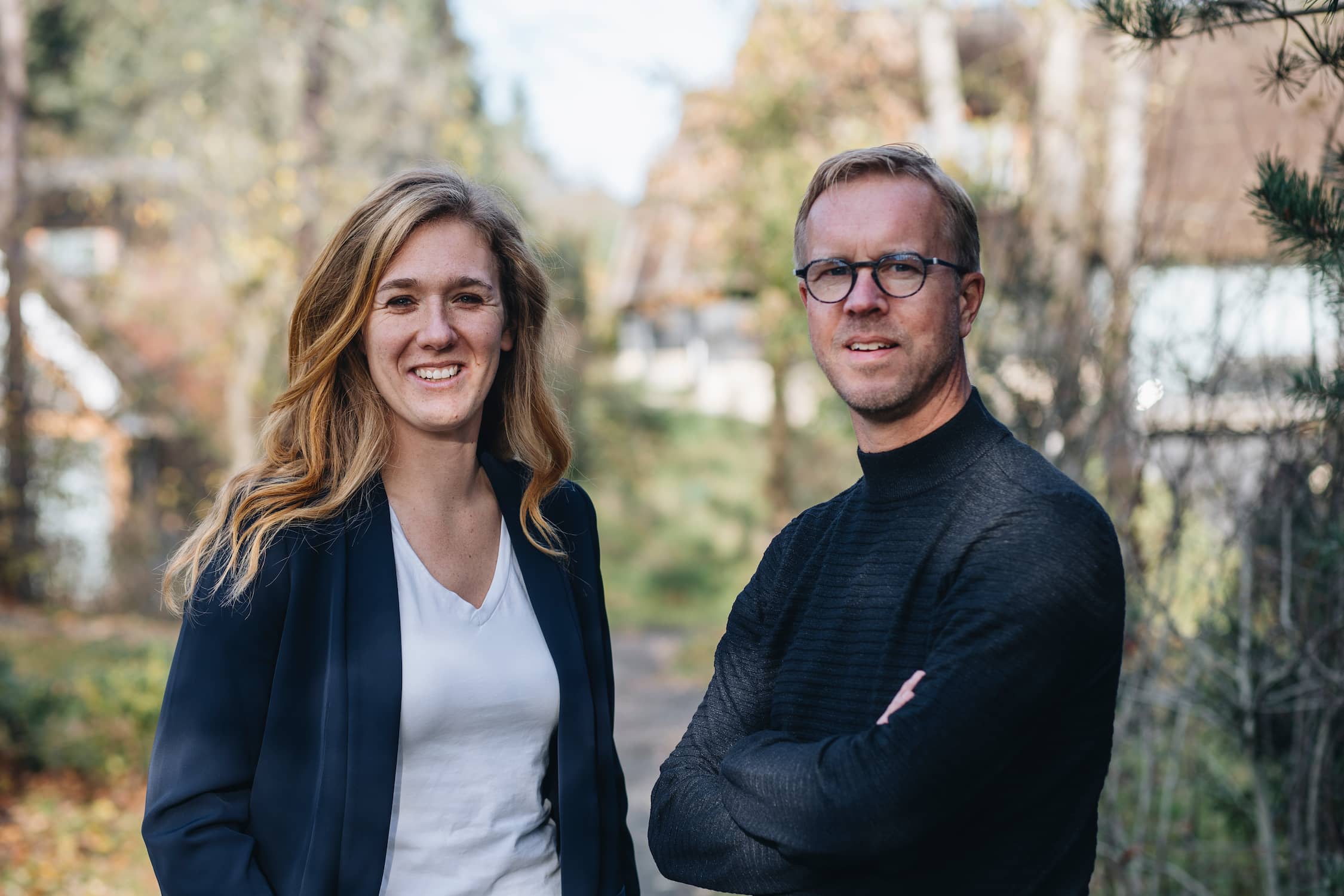 Twee volwassenen, een vrouw en een man met bril, buiten in een bosrijke omgeving met herfstbladeren.