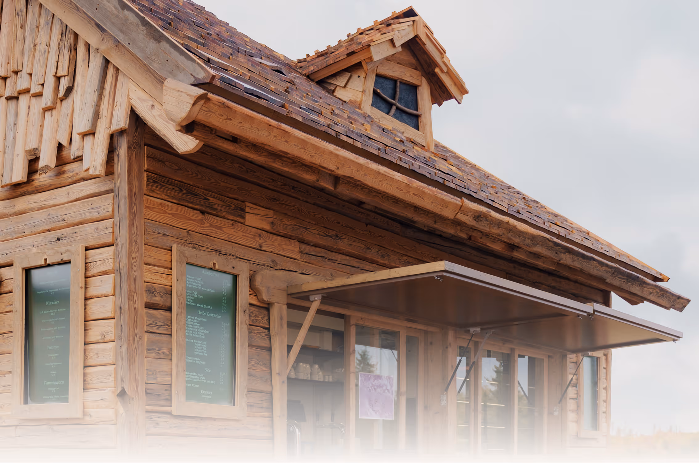 Holzhütte mit Schindeldach und offenem Fenster mit Markise, an der Außenwand sind grüne Speisekarten angebracht.