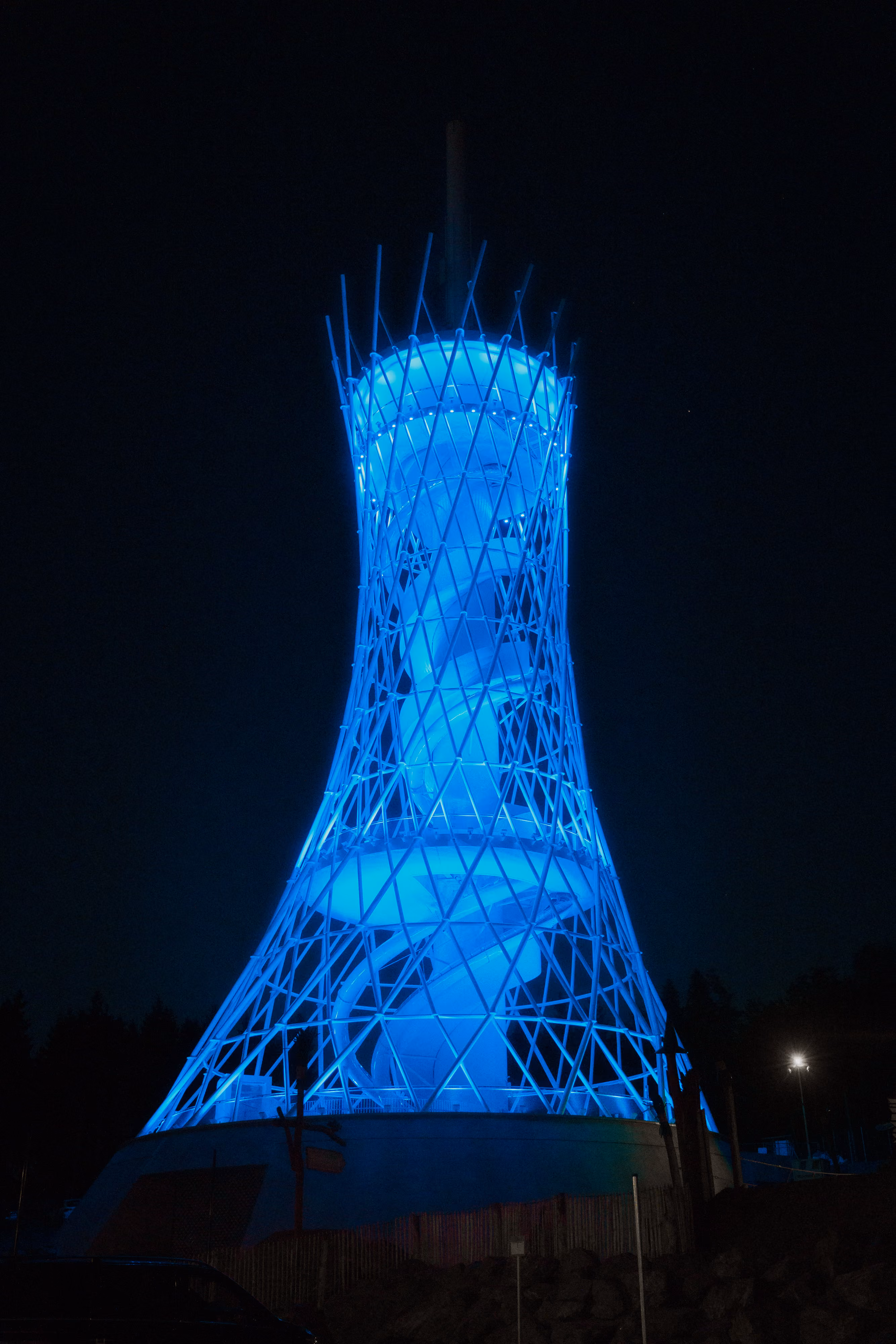 Beleuchteter, blauer HEX Erlebnisturm mit spiralförmiger Rutsche bei Nacht vor dunklem Himmel.