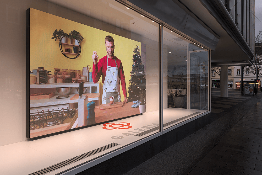 A large LED wall hangs in a shop window. It shows a man preparing a dessert.