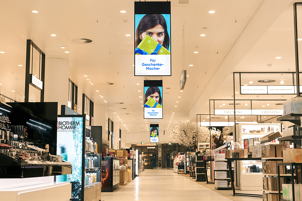 Cosmetics department of a department store with large screens hanging from the ceiling that display digital signage advertising.