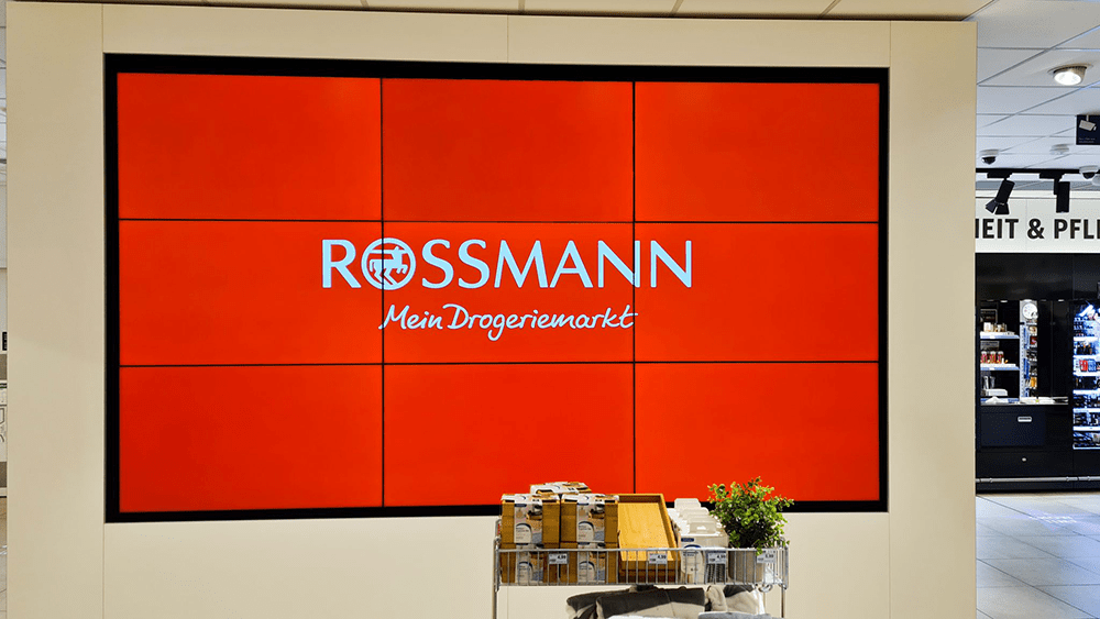 Eine große Digital Signage Wand, die aus 9 einzelnen LCD-Bildschirmen besteht. Sie zweigt das Logo der Drogeriemarktkette Rossmann in weiß auf rot.