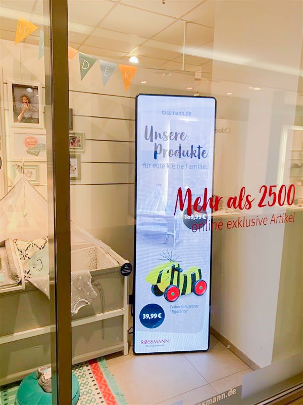 Ein LED Post steht in einem Schaufenster, das mit einem Kinderbettchen und Babyartikeln dekoriert ist. Das LED Poster spielt zum Thema passende Werbung aus.