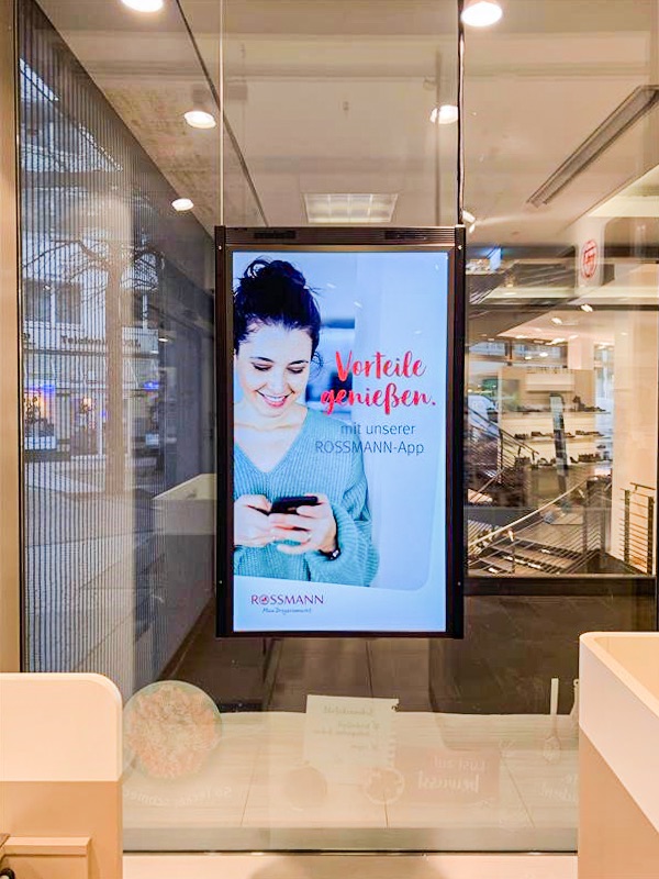 Ein LCD Bildschirm hängt von der Decke ab im Schaufenster eines Drogeriemarkts. Er spielt Digital Signage Werbung aus.