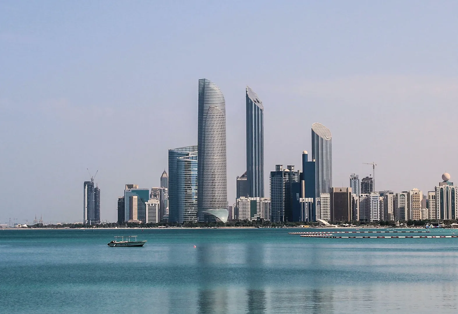 Skyline of Abu Dhabi with modern skyscrapers along a calm turquoise waterfront under a clear sky.