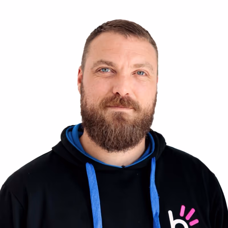 Man with a beard wearing a black hoodie with blue drawstrings and a logo, standing against a white background.