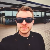 Man wearing sunglasses and a dark shirt standing outdoors on a wooden deck with a modern building in the background.