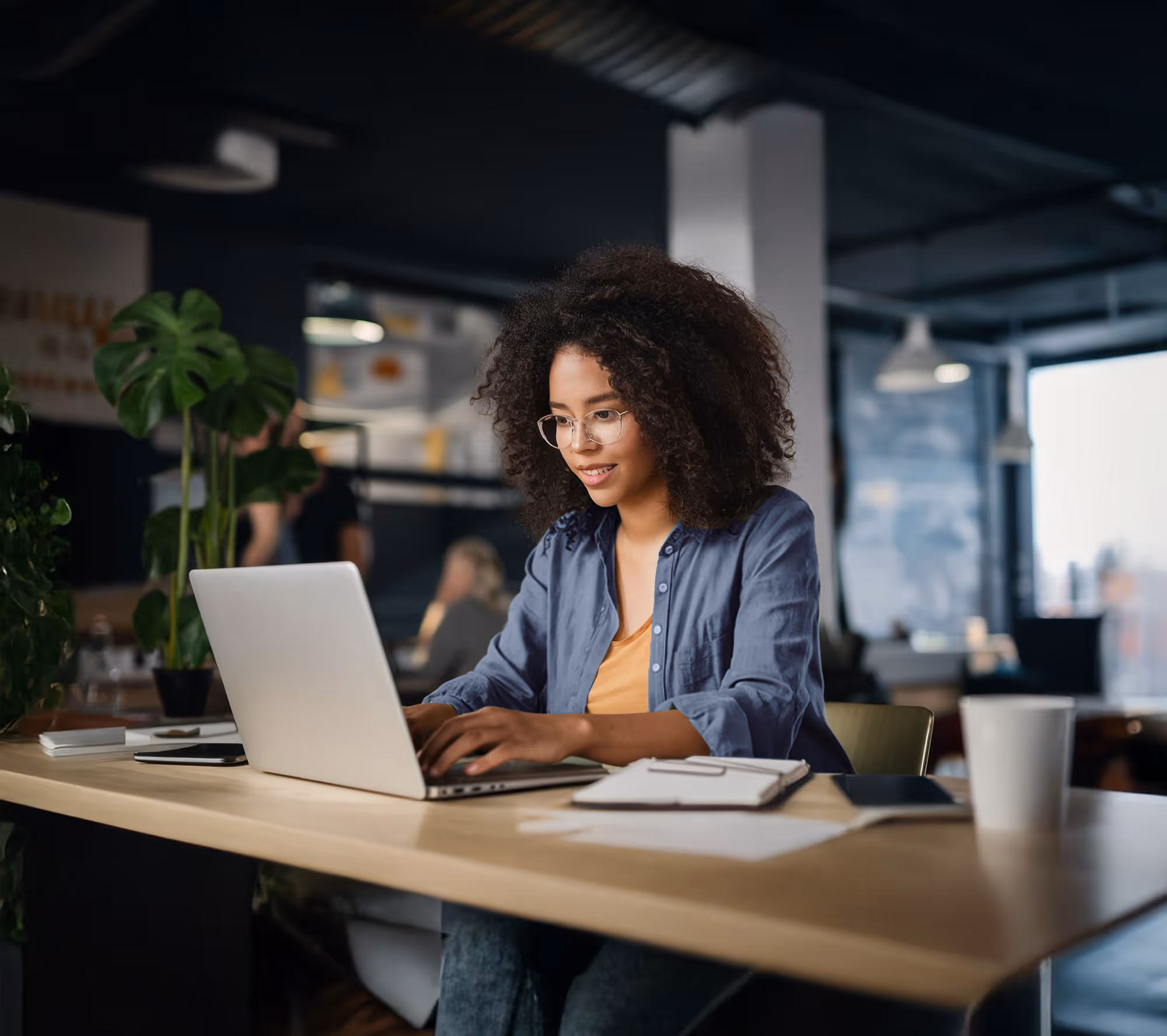 Smiling woman working on a laptop in a modern office space with plants and warm lighting.