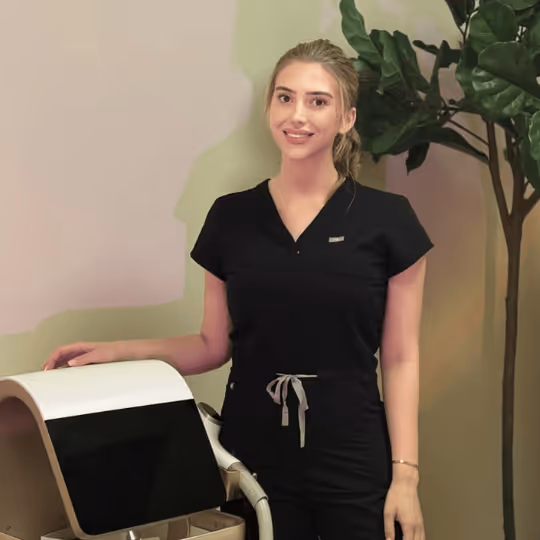 Smiling woman in black medical scrubs standing next to a medical device in a room with a plant at Better Med Spa