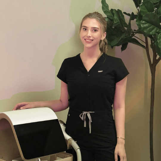 Smiling woman in black medical scrubs standing next to a medical device in a room with a plant.