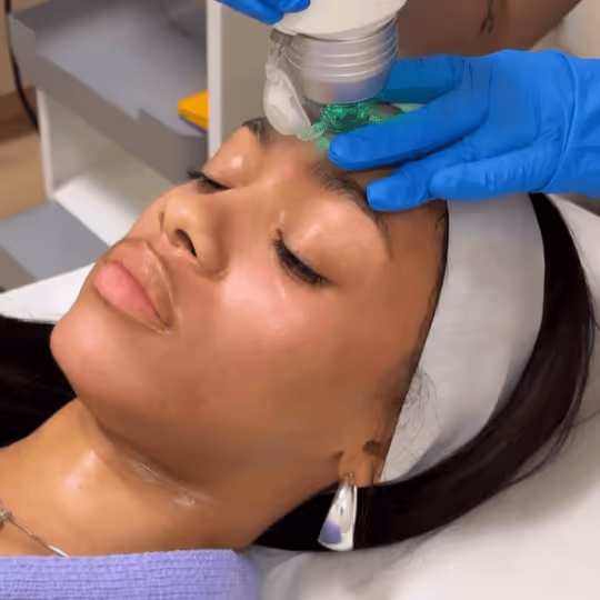 Woman receiving a facial treatment with a handheld device applied to her forehead by a person wearing blue gloves.