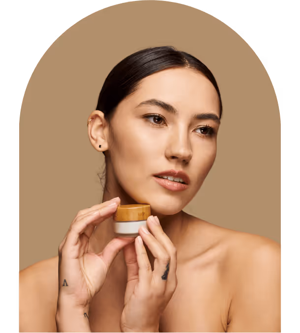 Woman with clear skin holding a small jar of cream with a wooden lid against a beige background.