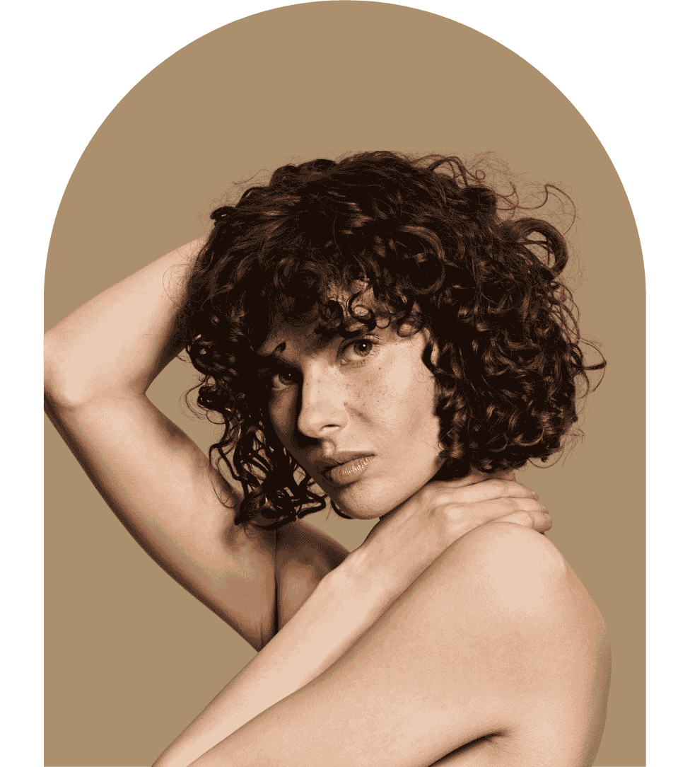 Portrait of a woman with curly dark hair looking at the camera, resting one hand on her neck and the other behind her head against a brown background.