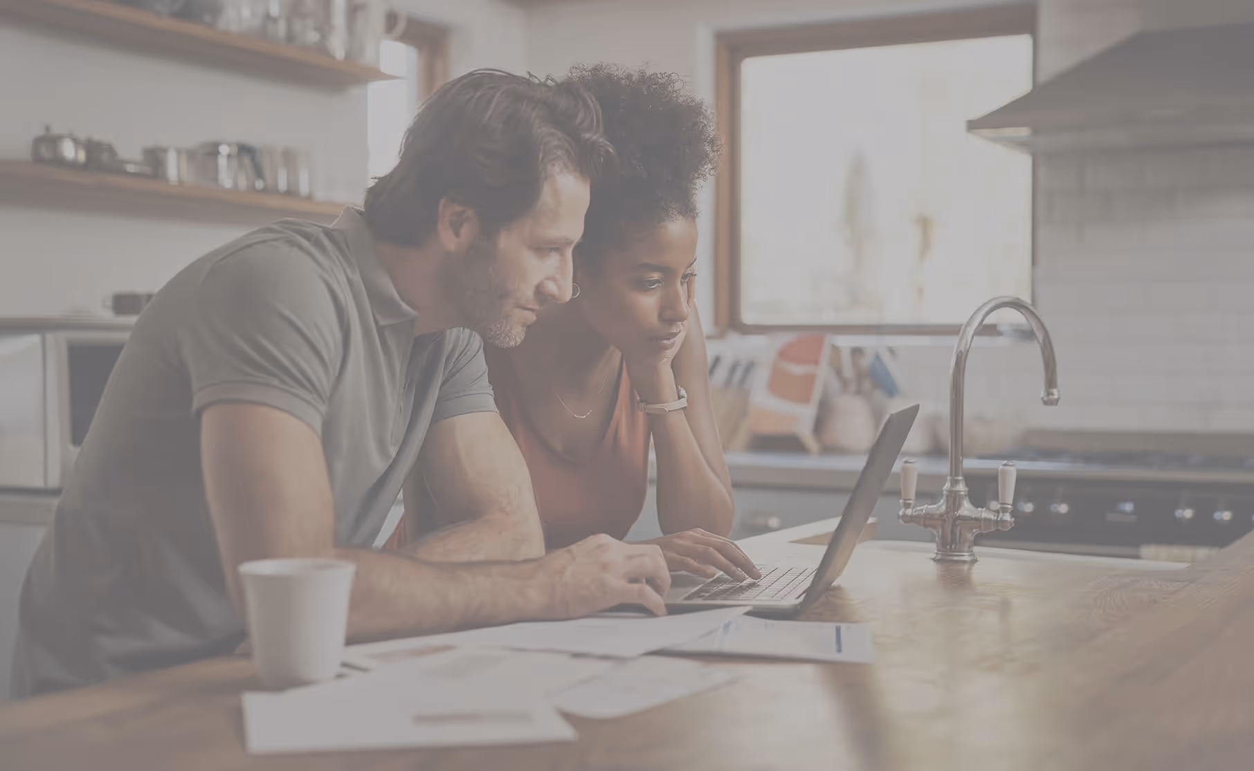 Man and woman sitting at a kitchen counter looking at a laptop together with papers and a coffee cup nearby.
