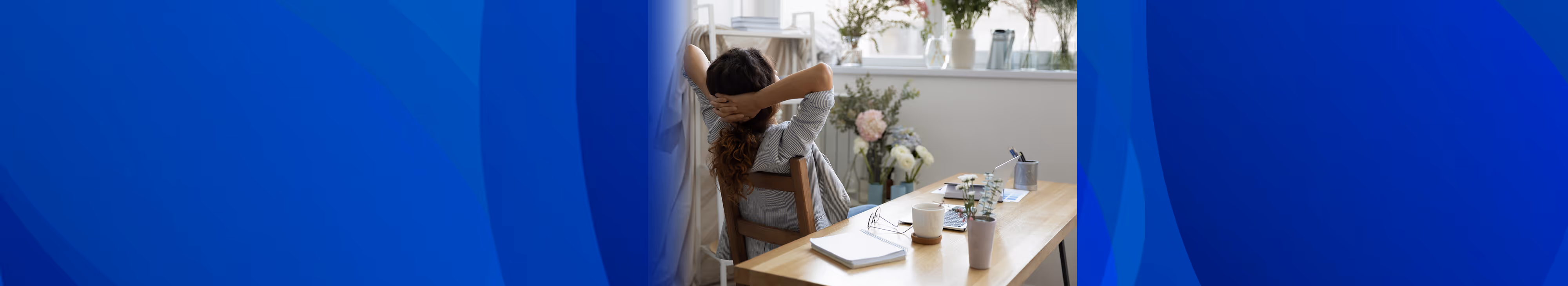 A woman sitting at a wooden desk in a bright room, stretching with hands behind her head, surrounded by notebooks, a cup, and plants.