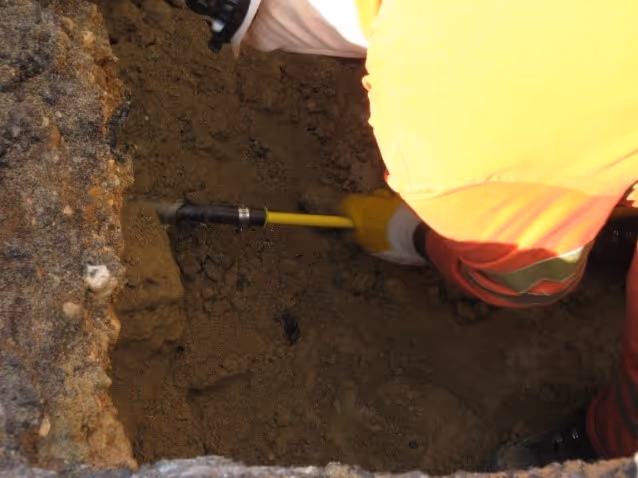Worker in yellow gloves and orange safety jacket inserting a yellow-handled tool into soil inside a dug hole.