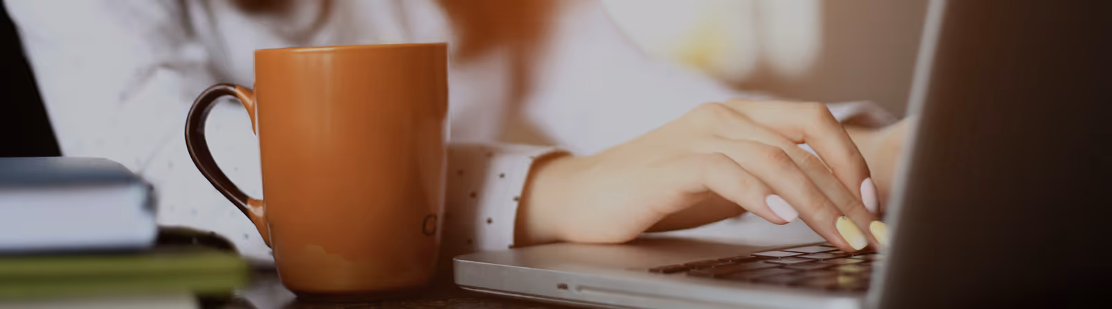 Person typing on a laptop with a brown coffee mug and stacked books nearby.