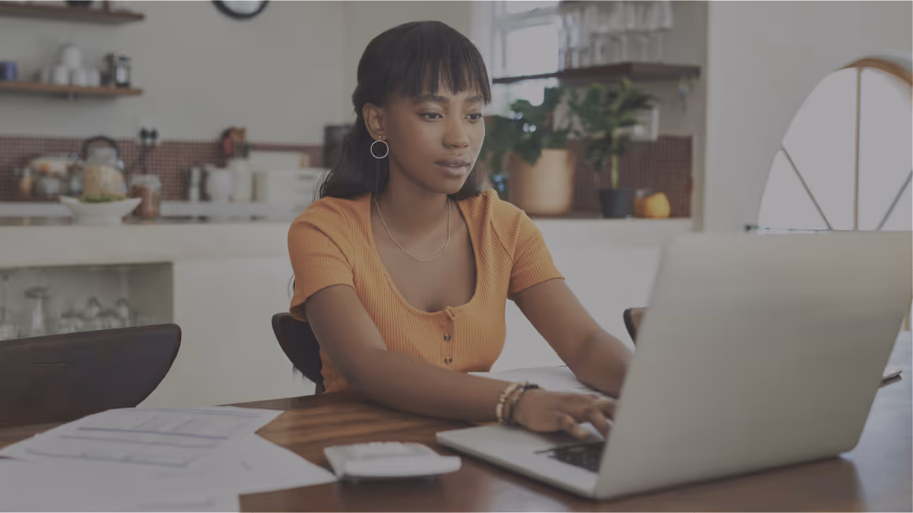 Woman with black hair in an orange top working on a laptop at a wooden table with papers and a phone.