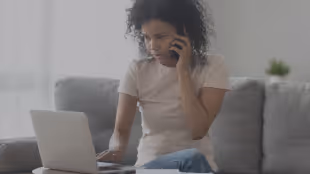Woman sitting on a couch using a laptop while talking on a smartphone.