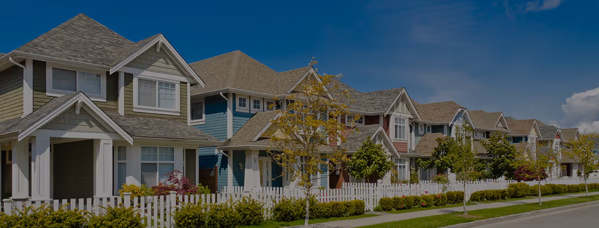Row of modern suburban houses with white fences, small trees, and blue sky in the background.