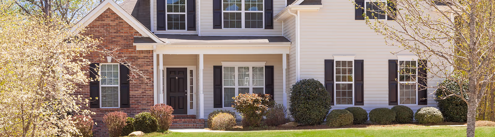 Front view of a suburban house with brick and white siding, black shutters, a black front door, and landscaped shrubs and trees.