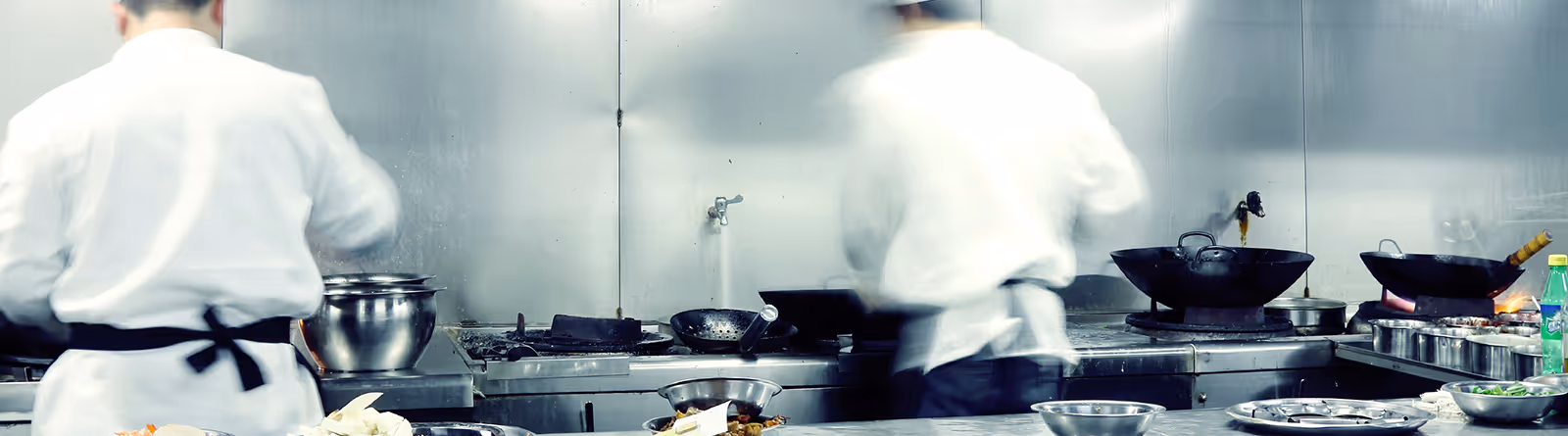 Two chefs wearing white uniforms working in a commercial kitchen with stainless steel counters and cooking utensils.