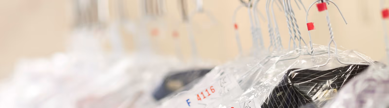 Close-up of several dry-cleaned clothes covered in plastic protective bags hanging on a rack with metal hangers.