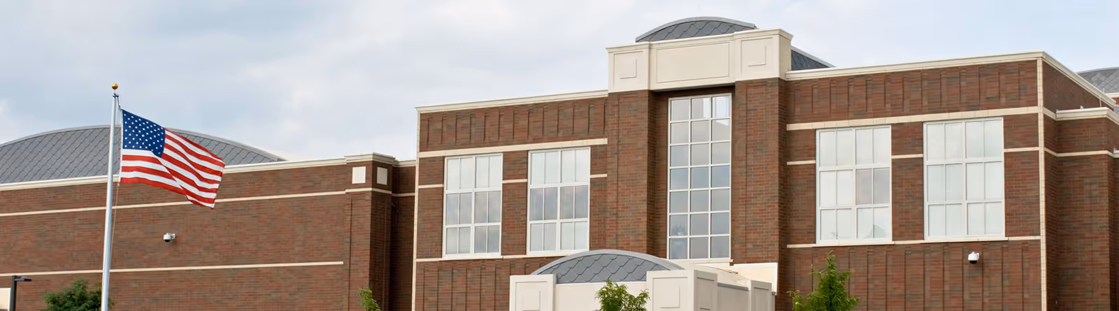 American flag waving in front of a large brick building with multiple windows and a gray roof.