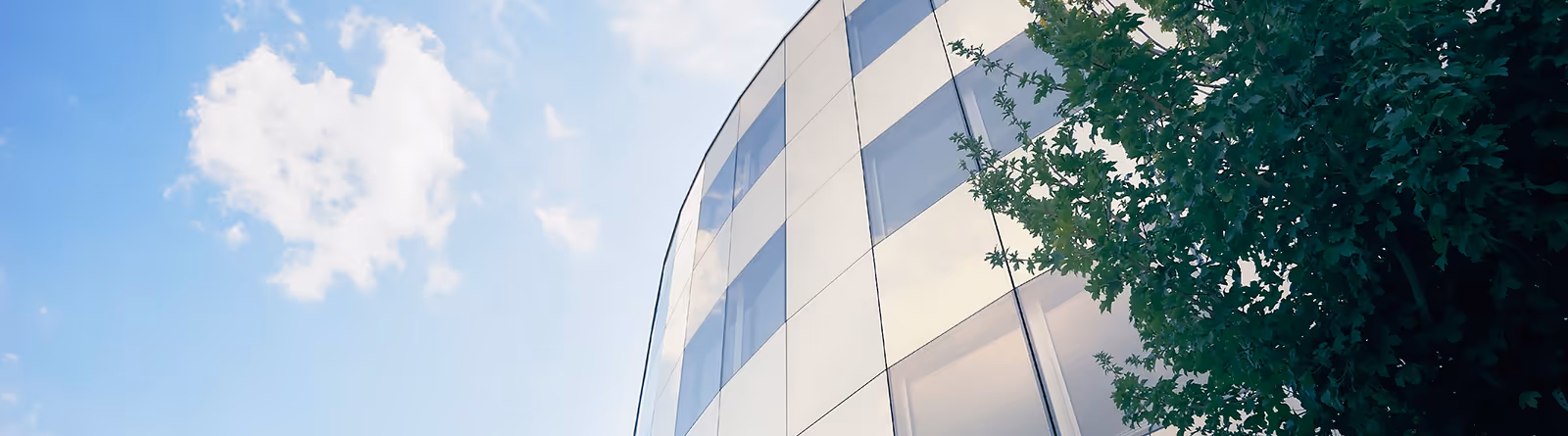 Curved modern building facade with glass windows next to green tree under a partly cloudy blue sky.