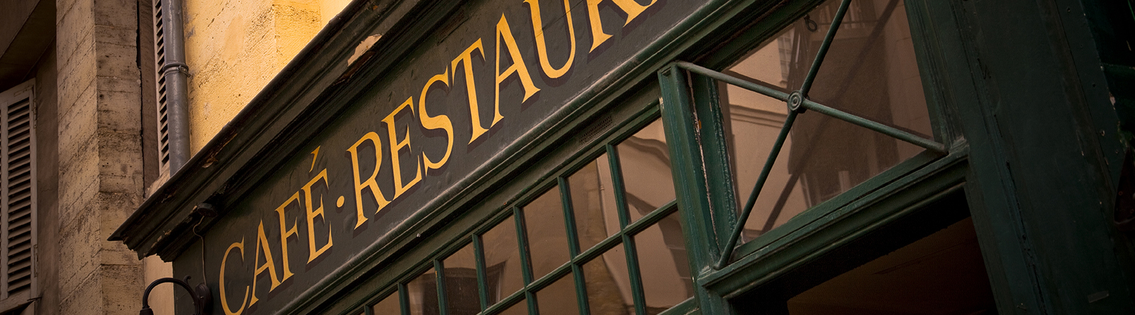 Exterior of a café-restaurant with yellow lettering on a green window frame reflecting nearby buildings.