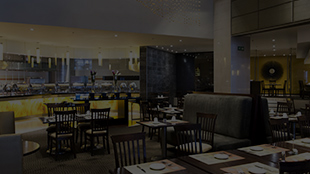 Dimly lit restaurant dining area with tables set for service and a counter with bar stools in the background.