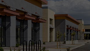 Exterior of a modern retail shopping center with storefronts and sidewalk under a cloudy sky.